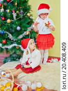 Two little girls dressed in white blouses, red skirts and santa caps in room with decorated christmas tree. Стоковое фото, фотограф Losevsky Pavel / Фотобанк Лори