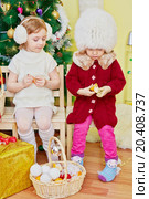 Two little girls sit on wooden chairs in room with decorated christmas tree and skin tangerin. Стоковое фото, фотограф Losevsky Pavel / Фотобанк Лори