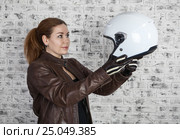 Woman motorcyclist wants to try white open helmet, holding in stretching arms. Brick wall background. Стоковое фото, фотограф Кекяляйнен Андрей / Фотобанк Лори