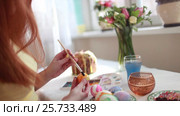 Redhead girl paints Easter eggs with a brush. on the table Easter cake, eggs, sweets, bouquet of flowers. Стоковое видео, видеограф Dmitry Chapurin / Фотобанк Лори