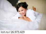 Joyful bride in the morning on the bed in bathrobe smiling cheerful, positive, with a ready hairdo and make-up, a wedding dress in the background. In anticipation of marriage. Стоковое фото, фотограф Евгений Майнагашев / Фотобанк Лори