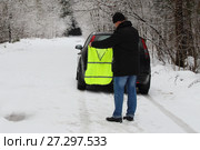 Мужчина водитель надевает световозвращающий жилет вне населенного пункта в условиях недостаточной видимости на заснеженной дороге зимой на фоне леса и автомобиля (2017 год). Редакционное фото, фотограф Илья Илмарин / Фотобанк Лори