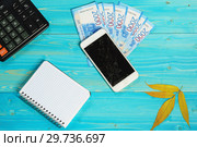Калькулятор, блокнот, смартфон с разбитым экраном и четырнадцать тысяч рублей новыми купюрами лежат на синем деревянном столе. Подсчет расходов на новый гаджет. Стоковое фото, фотограф Наталья Гармашева / Фотобанк Лори