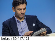 Министр РФ Михаил Абызов сидит с планшетом в президиуме конференции в городе Сочи, Россия (2017 год). Редакционное фото, фотограф Николай Винокуров / Фотобанк Лори