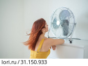 Portrait of a beautiful red-haired woman in a mustard jumpsuit enjoying the cooling breeze from a large electric fan. A smiling girl freshens up in the hot summer heat. Hair develops from the wind. Стоковое фото, фотограф Михаил Решетников / Фотобанк Лори