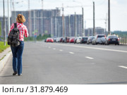 Rear view at Caucasian female hitchhiker walking on asphalt road in urban street, copy space. Стоковое фото, фотограф Кекяляйнен Андрей / Фотобанк Лори