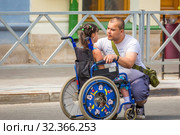 Russia, Samara, July 2019: Little sick disabled child in a wheelchair surrounded by father on a city street. Редакционное фото, фотограф Акиньшин Владимир / Фотобанк Лори
