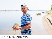 Только что пойманного большого сазана рыбак держит в руках на фоне реки (2019 год). Редакционное фото, фотограф Игорь Низов / Фотобанк Лори