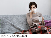 Sick woman suspected of covid-19, being at home in bed, measures the temperature and sneezes. Sick woman sitting under the blanket. Girl is quarantined with suspected coronavirus. Стоковое фото, фотограф Сергей Тимофеев / Фотобанк Лори