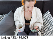 Stay at home. Social distancing. Woman at home relaxing on sofa couch drinking tea from white cup, listening to relaxing music, stay connected to friens and family via social networks on mobile. Стоковое фото, фотограф Matej Kastelic / Фотобанк Лори