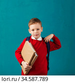 Cheerful thoughtful little school boy in school uniform with backpack and big pile of books standing against blue wall. Looking at camera. Back to School. Стоковое фото, фотограф Nataliia Zhekova / Фотобанк Лори