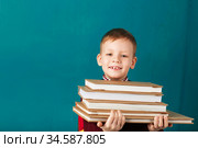 Cheerful thoughtful little school boy in school uniform with backpack and big pile of books standing against blue wall. Looking at camera. School concept. Стоковое фото, фотограф Nataliia Zhekova / Фотобанк Лори