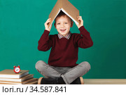 Cheerful smiling little school boy with big heavy books on his head sitting on a desk against turquoise wall. Looking at camera. School concept. Стоковое фото, фотограф Nataliia Zhekova / Фотобанк Лори