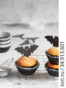 Halloween pumpkin muffins in black capsules decorated with cardboard bats. Festive Halloween cupcakes. Close-up view of delicious spooky halloween muffins on the grey table. Halloween recipes. Стоковое фото, фотограф Nataliia Zhekova / Фотобанк Лори