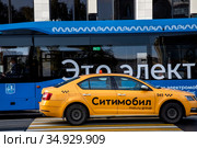 Желтое такси с надписью "Ситимобил" на дверях автомобиля на фоне электробуса в центре города Москвы, Россия (2020 год). Редакционное фото, фотограф Николай Винокуров / Фотобанк Лори