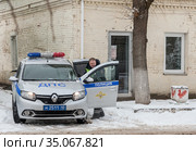 Таруса. Сотрудник полиции стоит у стены дома рядом с распахнутой дверцей служебного автомобиля (2020 год). Редакционное фото, фотограф Наталья Николаева / Фотобанк Лори