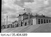 Несколько человек в форме стоят вблизи здания Речного вокзала, белое двухэтажное строение с флагом на крыше, территория гавани в летнюю погоду, Осетрово, Чукотка, 1963 год. Стоковое фото, фотограф Варфоломей Тетерин / Фотобанк Лори