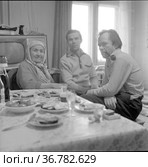 Двое мужчин и пожилая женщина позируют для фотопортрета, они сидят за столом с едой и смотрят в камеру, телевизор и окно на заднем плане , Токарев и Морозов в гостях у Тетериных. Редакционное фото, фотограф Варфоломей Тетерин / Фотобанк Лори