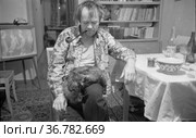 Мужчина курит трубку с табаком во время застолья, он сидит в расстёгнутой рубашке и котом на коленях, человек рядом со столом, шкафом и телевизором , Токарев и Морозов в гостях у Тетериных. Редакционное фото, фотограф Варфоломей Тетерин / Фотобанк Лори