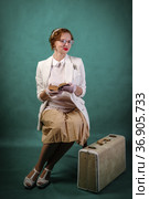 Young woman with glasses, dressed in retro style, sitting on a chair, with a suitcase at her feet, with a book in her hands. Studio portrait. Стоковое фото, фотограф Вадим Орлов / Фотобанк Лори