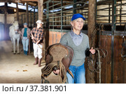 Elderly female rancher walking in stable with saddle in hands. Стоковое фото, фотограф Яков Филимонов / Фотобанк Лори