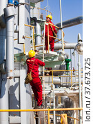 Two Asian maintenance workers in yellow hardhats and red work wear on distillation column of oil refinery plant on blue sky background. CNPC-AMG. company. (2012 год). Редакционное фото, фотограф Alexey Rezvykh / Фотобанк Лори