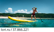 Tanned man stands on a yellow supboard and paddles. Active sport on the beach in the turquoise sea water. Стоковое фото, фотограф Енваев Андрей / Фотобанк Лори