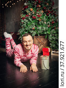 Cheerful funny man in pink sleepwear lying down near decorated fir tree and Christmas present. Стоковое фото, фотограф Darkbird77 / Фотобанк Лори