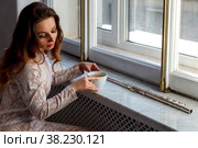 At window sits a girl-flutist with a Cup of tea. The flute is on windowsill. With space on photo for your lettering or logo. Стоковое фото, фотограф Aleksandr Golubev / Фотобанк Лори