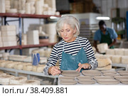 Portrait of elderly woman in apron cutting burrs off freshly made earthenware cups and plates with knife. Стоковое фото, фотограф Яков Филимонов / Фотобанк Лори