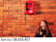 Funny little girl in leather jacket looking on old red payphone at brick wall backdrop. Small brunette lady model looking up on telephone. Communication concept. Copy space for advertising text. Стоковое фото, фотограф Aleksandr Golubev / Фотобанк Лори