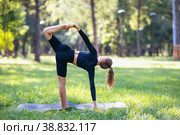Девушка практикующая йога выполняет вариацию упражнения Ардха Чандрасана, позу полумесяца, тренируется в парке теплым летним утром стоя на коврике. Стоковое фото, фотограф Сергей Чайко / Фотобанк Лори