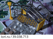 medicines and pills are lying in a small shopping cart against the background of money euro dollars 100 500. pills are poured out of the cart for money the problem of the cost of medicines expensive m. Стоковое фото, фотограф Meshkov Sergey / Фотобанк Лори