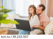 Happy family, daughter sitting on her father's lap on couch at home looking at laptop monitor together. Стоковое фото, фотограф Мария Мороз / Фотобанк Лори