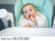 A hungry baby is gnawing on a plastic spoon at the table on a high chair. Teething, whims, itchy gums, introduction of complementary foods. Стоковое фото, фотограф Ольга Симонова / Фотобанк Лори