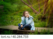 Восьмилетняя девочка наблюдает за природными явлениями под лучами солнца в лесу. Ребенок сидит на мосту, скрестив ноги. Стоковое фото, фотограф Юлия Мелешина / Фотобанк Лори
