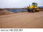 Ust-Luga, Leningrad oblast, Russia - November 16, 2021: Asphalt compactor Bomag flatten sand subbase Road construction site. Chemical waste pit on background. Редакционное фото, фотограф Alexey Rezvykh / Фотобанк Лори