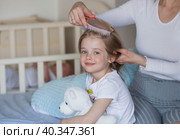 A happy young mother combs her adorable little daughter's hair while sitting in bed in the morning. The girl smiles. Family and care. Стоковое фото, фотограф Photojuli / Фотобанк Лори