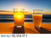 A glass of orange juice on the sea sand, small waves, romantic sunrise. Стоковое AI-изображение, AI-художник Сергей Елизарьев / Фотобанк Лори