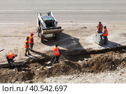 ANGARSK, IRKUTSK REGION, RUSSIA - May 18, 2023: Workers in signal red vests lay a curb stone on the side of a road being repaired. Mini tractor loader brings soil in the bucket. Редакционное фото, фотограф Виктория Катьянова / Фотобанк Лори