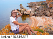 A woman with a scythe sits on a stone and squints from the sun against the backdrop of a beautiful sea rocky landscape with a sandy beach, top view. Travel and tourism. Стоковое фото, фотограф Коханова Наталья / Фотобанк Лори