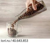 Кофейные зерна высыпают из прозрачной стеклянной бутылки в прозрачную стеклянную кружку. Coffee grains in a glass bottle and a glass on a wooden background. Стоковое фото, фотограф Валерий Елистратов / Фотобанк Лори