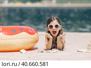 Surprised emotional cute little girl in sunglasses lying on the sand on a beach in a swimsuit near a straw hat. Стоковое фото, фотограф Ирина Михайличенко / Фотобанк Лори