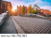 Arch bridge over canals with cobbled road in the Speicherstadt of Hamburg, Germany, Europe. Historical red brick building lit by golden sunset light near water castle palace (2019 год). Стоковое фото, фотограф Igor Tichonow / Фотобанк Лори