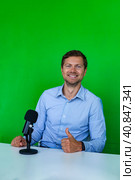 Smiling handsome man broadcaster with microphone at desk thumb up on green backdrop, looking at camera. Male broadcaster sitting in television studio. Tv production concept. Copy ad text space banner. Стоковое фото, фотограф Aleksandr Golubev / Фотобанк Лори