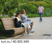 Moscow, Russia - May 20, 2021: Young pretty women sitting and resting on wooden bench in the public park. Hot spring day. Everybody use smartphones. Youth leisure concept. Walking man in distance. Редакционное фото, фотограф YuryKara / Фотобанк Лори