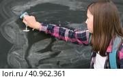 Teenage student girl in shirt with blue backpack writing with chalk numbers and letters on black chalkboard background. Education, back to school and people concept. Joyful Teen girl near blackboard. Стоковое видео, видеограф Марина Демидюк / Фотобанк Лори
