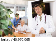positive medical male colleague standing in meeting room and employees are talking behind him at table. Стоковое фото, фотограф Яков Филимонов / Фотобанк Лори