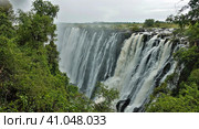 The powerful streams of Victoria Falls plunge into the abyss. The rocky channel of the Zambezi River is visible. In the foreground, green tropical vegetation. Zambia (2020 год). Стоковое фото, фотограф Вера Ивановна Кубарева / Фотобанк Лори