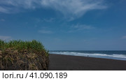 A beach with black volcanic sand on the Pacific coast. Turquoise surf waves roll onto the shore. A tiny silhouette of a man is visible. Blue sky. There is green grass in the foreground. Стоковое фото, фотограф Вера Ивановна Кубарева / Фотобанк Лори